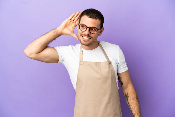 Camarero de restaurante brasileño sobre fondo púrpura aislado escuchando algo poniendo la mano en la oreja - Foto, Imagen
