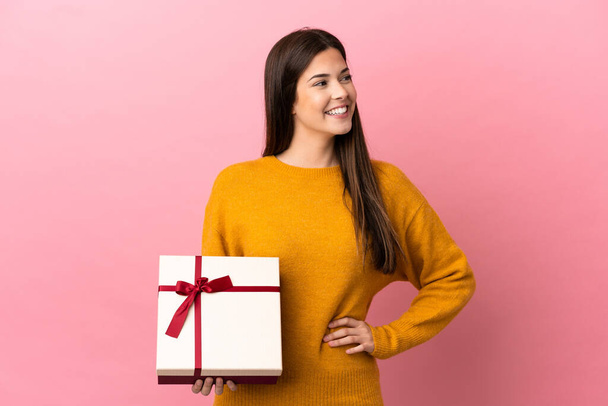 Teenager Brazilian girl holding a gift over isolated pink background posing with arms at hip and smiling - Foto, immagini