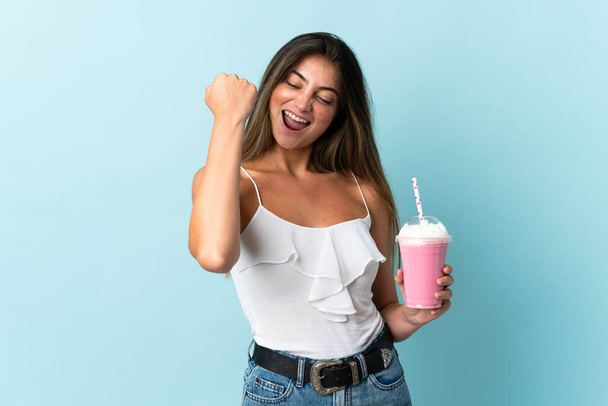 jonge vrouw met aardbei milkshake geïsoleerd op blauwe achtergrond vieren een overwinning - Foto, afbeelding