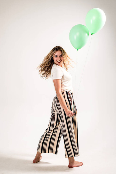 A girl with wavy blonde hair in a light shirt and striped pants with blue balloons in her hand, having fun waiting for a holiday in her honor. Isolated on a white background. - Fotografie, Obrázek