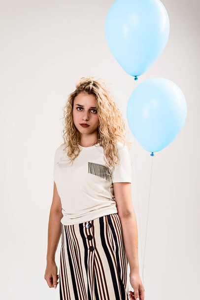 A girl with wavy blonde hair in a light shirt and striped pants with blue balloons in her hand, sad not waiting for the holiday in her honor. Isolated on a white background. - Fotoğraf, Görsel