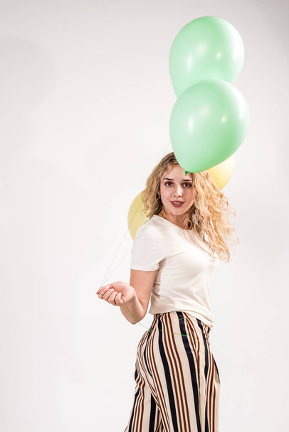 Смішна дівчина, блондинка, в легкій футболці і смугастій штани-спідниці. дівчина стоїть босоніж з повітряними кулями в руках на білому тлі
. - Фото, зображення