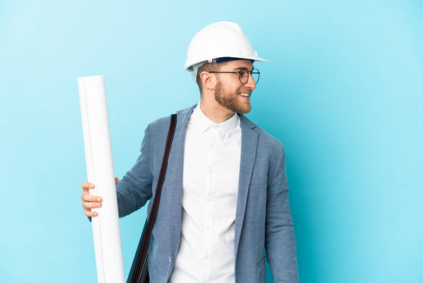 Jeune architecte homme avec casque et tenant des plans sur fond isolé regardant côté - Photo, image