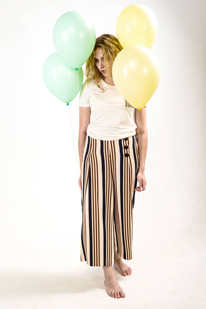 Sad girl, blonde, in a light T-shirt and striped trousers-skirt. a girl stands barefoot with balloons in her hands on a white background. - 写真・画像