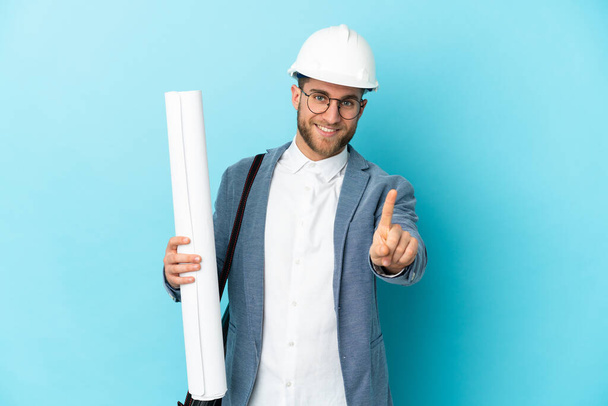 Jeune architecte avec casque et tenant des plans sur fond isolé montrant et levant un doigt - Photo, image