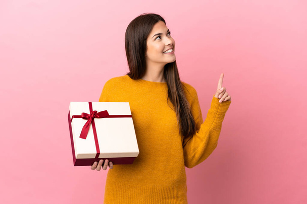 Teenager Brazilian girl holding a gift over isolated pink background pointing up a great idea - Φωτογραφία, εικόνα