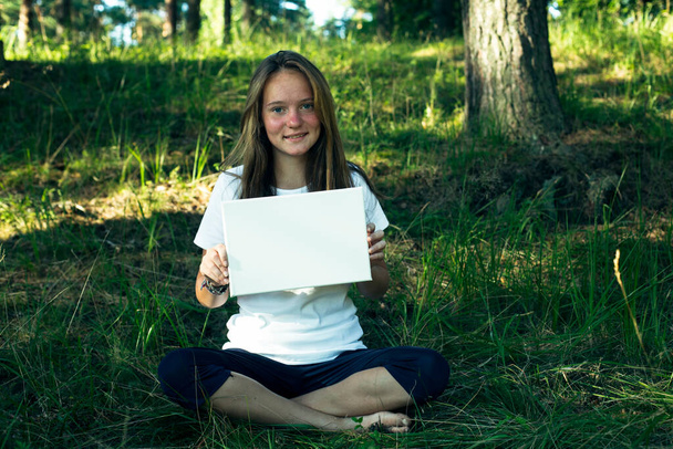A girl holding clean white sheet paper (banner for message), in the green park. - Photo, Image
