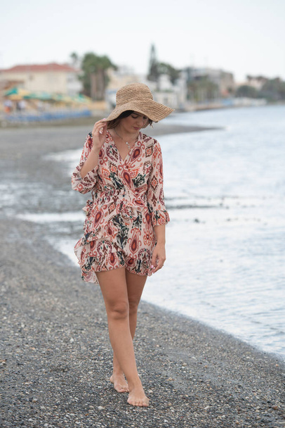 Jeune femme caucasienne portant une robe d'été élégante et un chapeau marchant sur la plage. Concept d'été à la plage - Photo, image