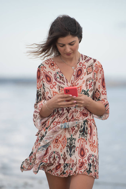 Sonriente joven morena caucásica mujer charlando en un teléfono móvil rojo en la playa. Concepto de vida - Foto, imagen