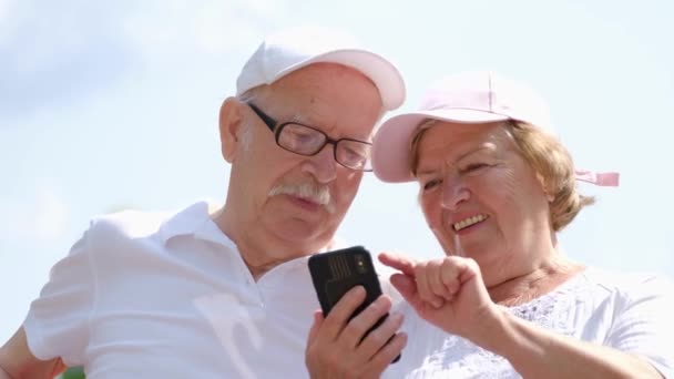 Современные бабушки и дедушки используют смартфон, им весело сидеть в городском парке - Кадры, видео
