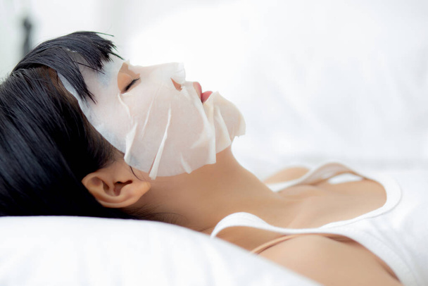 Beautiful young asian woman with sheet facial mask and sleep on bed at bedroom, beauty girl applying cosmetic and makeup with skin care face for wrinkle, skincare treatment, health and wellness. - Фото, изображение