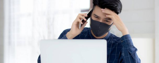 Yüz maskeli Asyalı genç bir iş adamı akıllı telefondan konuşuyor ve bilgisayar başında Coperid-19 'u korumak için çalışıyor. İş adamı karantina işini evden defter ve sosyal mesafeyle yapıyor ve evde kalıyor.. - Fotoğraf, Görsel