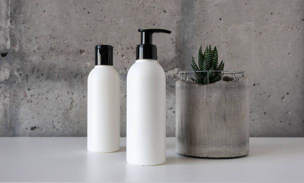 Botellas cosméticas y una suculenta maceta de hormigón sobre escritorio blanco sobre fondo de pared de cemento - Foto, imagen