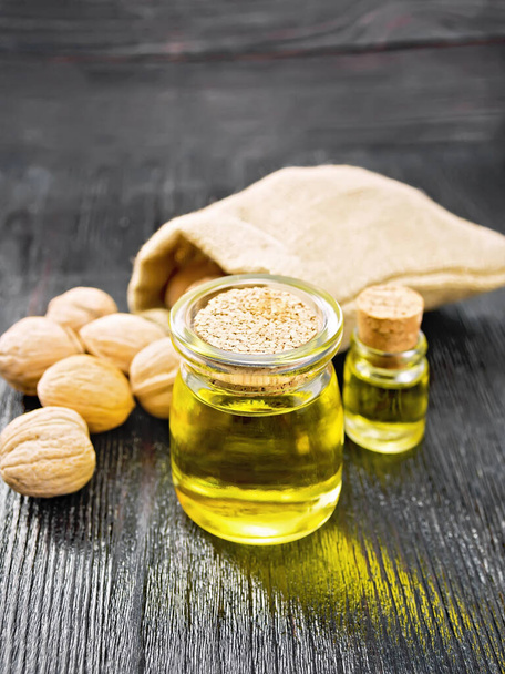 Walnut oil in a jar and a bottle, whole nuts in bag and on table on wooden board background - Photo, Image