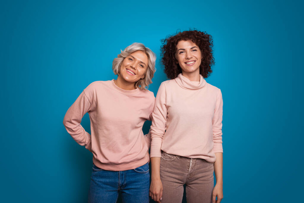 Dva přátelé pózují na modrém pozadí. Jedna dívka s blond vlasy, krátkými a rovnými, ale druhá s kudrnatými a hnědými vlasy - Fotografie, Obrázek