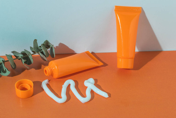 Gepresste Sahne, orangefarbene Tuben und ein Eukalyptuszweig auf orange-weißem Hintergrund. Kosmetische Cremes. - Foto, Bild