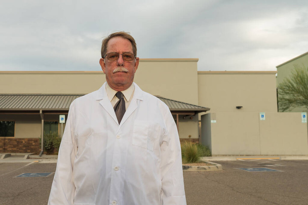 Starzejący się lekarz w białym fartuchu laboratoryjnym na zewnątrz przed kliniką. - Zdjęcie, obraz