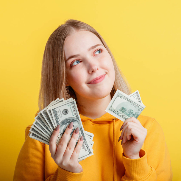 Happy teenage blonde girl holding in hands money cash dollars dreaming thoughtfully isolated on color yellow background. Portrait young excited smiling woman stack of money banknotes. Closeup square - Foto, Imagen