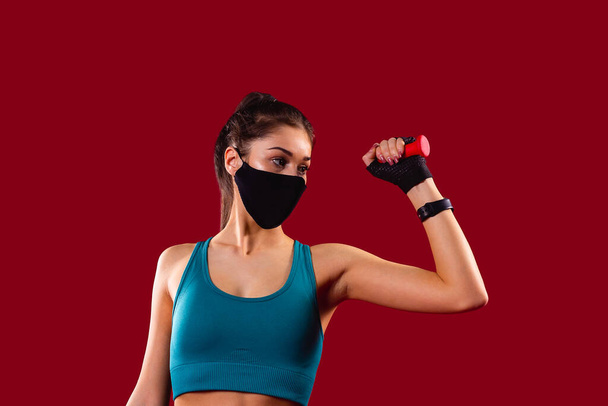Fitness girl with a textile mask posing in gym in blue top holding one red dumbbell in her hand raised it bending her arm in the log. - 写真・画像