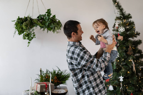 Family at Christmas at home, father plays with his son holding in his arms near the Christmas tree in the living room. New Year home holiday concept. - Photo, Image