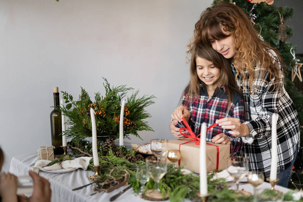 Glückliche Familienmütter und -töchter öffnen am festlich gedeckten Tisch eine weihnachtliche Geschenkbox. Heiligabend zu Hause feiern. Weihnachtliches Konzept. Hintergrund. - Foto, Bild