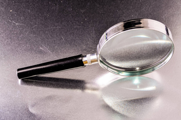 A close-up shot of a handheld magnifying glass with a metal frame reflected on a silver surface - Valokuva, kuva