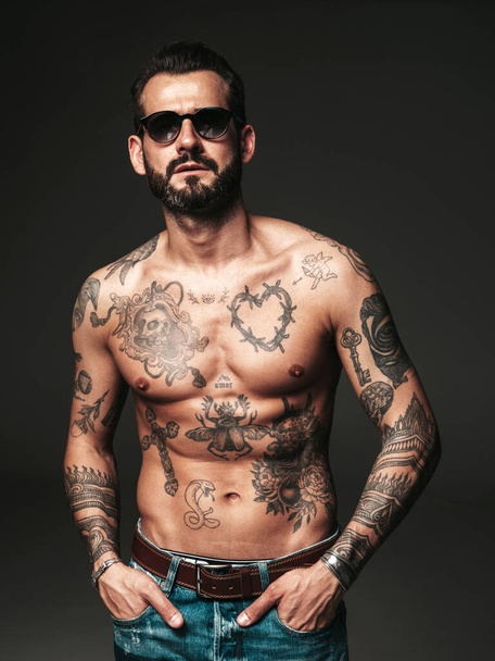 Portrait of handsome confident stylish hipster lambersexual model. Sexy modern man. Naked torso with tattoos.Fashion male posing in studio on dark background in sunglasses - Zdjęcie, obraz
