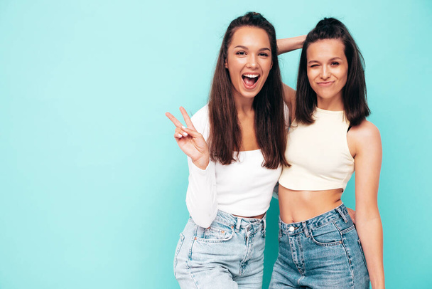 Two young beautiful smiling brunette hipster female in trendy summer clothes. Sexy carefree women posing near blue wall. Positive models having fun. Cheerful and happy. Show peace sign - Foto, imagen