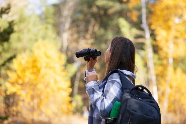 A young female tourist walks through the autumn forest, looking through binoculars - Фото, изображение
