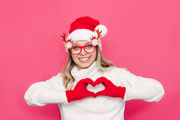 beautiful young woman in santa hat and glasses showing heart sign on pink background - Fotó, kép