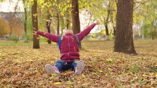 дівчина з рюкзаком кидає сухе осіннє листя над головою, шкільний мішок на плечах школярки, дитина в міській упаковці грає з жовтим листям, щаслива маленька дитяча природа на відкритому повітрі
 - Фото, зображення