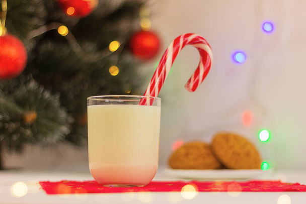 Weihnachtsmilchgetränk im Glas mit Zuckerrohr auf dem Hintergrund von Weihnachtsbaum und Lichtern. Traditioneller Leckerbissen für den Weihnachtsmann - Foto, Bild