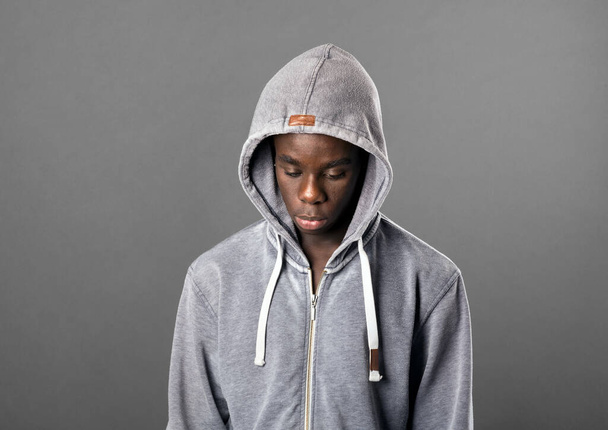 Young Black man wearing a grey hoodie looking down with a serious sombre expression in a close up head and shoulders portrait on grey - Foto, immagini