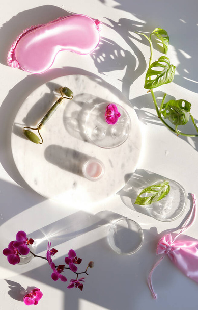 Jade face roller, moisturizing cream on marble plate and pink silk sleep mask. Exotic flat lay with monstera leaves and vibrant magenta orchid flowers. Sunlight, long shadows. Facial massage top view - Photo, image
