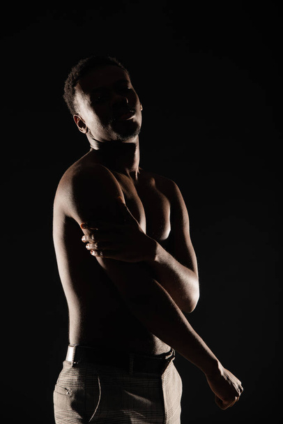 Joven macho negro posando con sus brazos sobre fondo oscuro, silueta - Foto, imagen