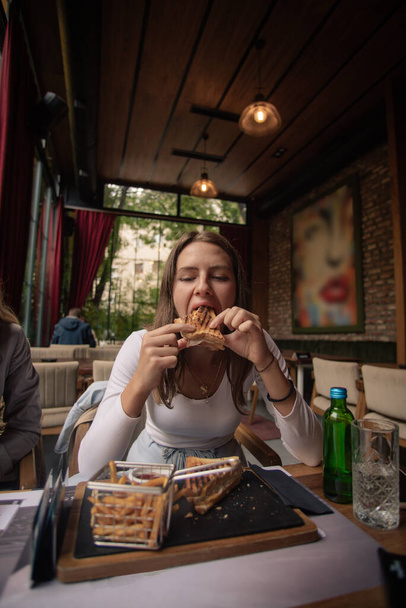 Η όμορφη και χαριτωμένη νεαρή κοπέλα τρώει μια μεγάλη μπουκιά από το σάντουιτς της στο μπαρ. - Φωτογραφία, εικόνα