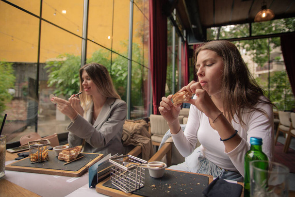 Una chica atractiva está comiendo en el restaurante y disfrutando de su comida, mientras que la otra está arreglando su maquillaje - Foto, imagen