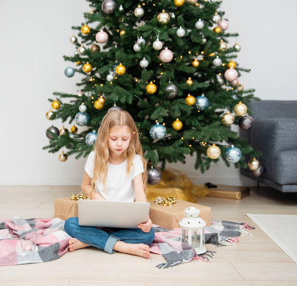 Χριστουγεννιάτικο δέντρο σε ένα μεγάλο σαλόνι. Το κοριτσάκι παίζει κοντά στο χριστουγεννιάτικο δέντρο. Ένα κορίτσι με φορητό υπολογιστή επικοινωνεί με φίλους. - Φωτογραφία, εικόνα