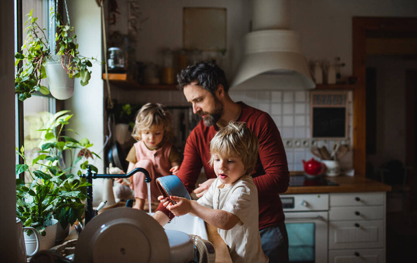 Mature father with two small children washing dishes indoors at home, daily chores concept. - Фото, изображение