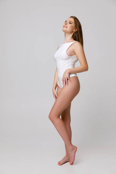 Full-length portrait of beautiful young woman in white bodysuit posing isolated over gray background - Valokuva, kuva