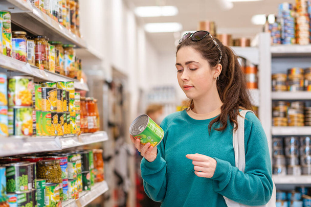 A young pretty Caucasian woman reads ingredients on a tin of canned peas. The concept of buying products and shopping. - Φωτογραφία, εικόνα