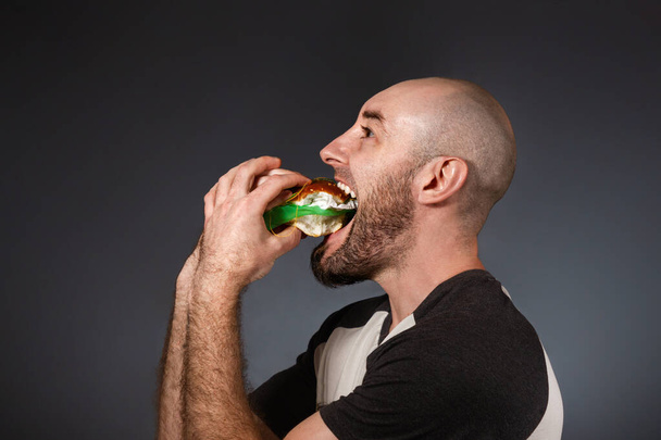 A gyorsétterem és az ökológia fogalma. Egy kopasz, szakállas férfi, aki boldogan eszik szeméttel töltött hamburgert. Egy groteszk, megnagyobbodott állkapocs. Fekete háttér. Fénymásolási hely. - Fotó, kép