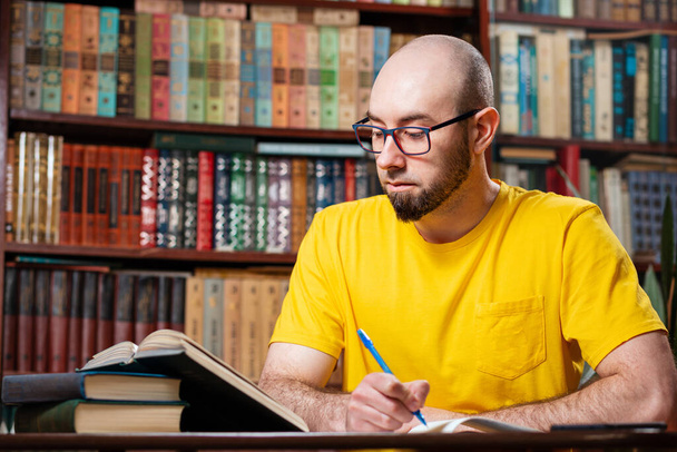 Un hombre calvo y barbudo con gafas está escribiendo algo en un cuaderno, mirando un libro. Hay estanterías en el fondo. Concepto de educación y aprendizaje a distancia. - Foto, imagen