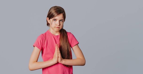 Portrait of cute young girl pulling lips down in upset smile, frowning and holding palms in pray while begging for apology, standing over gray background with copy free space for text - Photo, Image