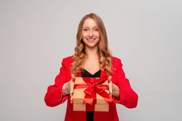 Sonriente joven rubia sosteniendo caja de regalo decorada con lazo rojo. Chica atractiva en elegante chaqueta roja posando aislado sobre fondo gris. Año Nuevo Día de la Mujer cumpleaños concepto de vacaciones - Foto, Imagen