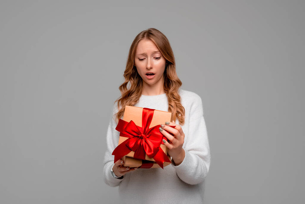 Eine junge Frau im weißen Pullover blickt mit schockiertem Gesichtsausdruck in die Geschenkbox, packt heimlich ihr Geschenk aus und steht vor grauem Hintergrund. Neujahrsempfang zum Frauentag - Foto, Bild