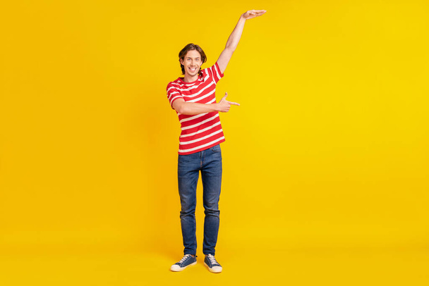 Photo of funny charming young guy dressed red t-shirt pointing finger showing empty space height smiling isolated yellow color background - Foto, Imagen