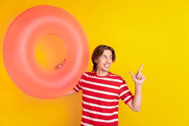 Photo of positive cute young guy dressed red t-shirt holding bloated circle pointing looking empty space isolated yellow color background - Foto, Imagen