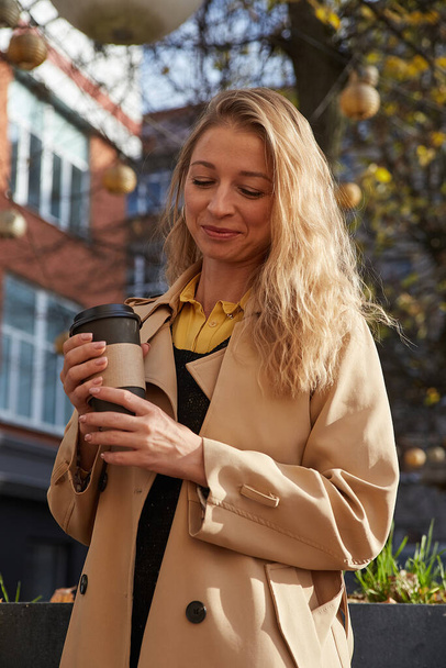Kaukasische lachende vrouw in beige trench jas met kopje koffie buiten - Foto, afbeelding