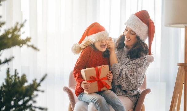 Merry Christmas, Happy Holidays. Cheerful mom and her cute daughter girl exchanging gifts. Parent and little child having fun near tree indoors.   - Fotoğraf, Görsel
